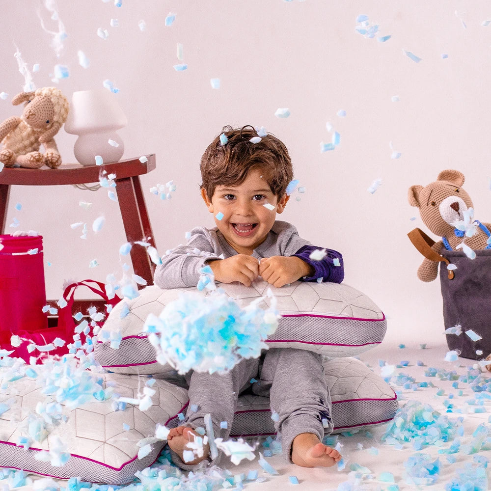 Criança sorridente brincando com travesseiros infantis snow kids, rodeada por flocos de espuma memory foam em um ambiente alegre e descontraído, com brinquedos ao fundo