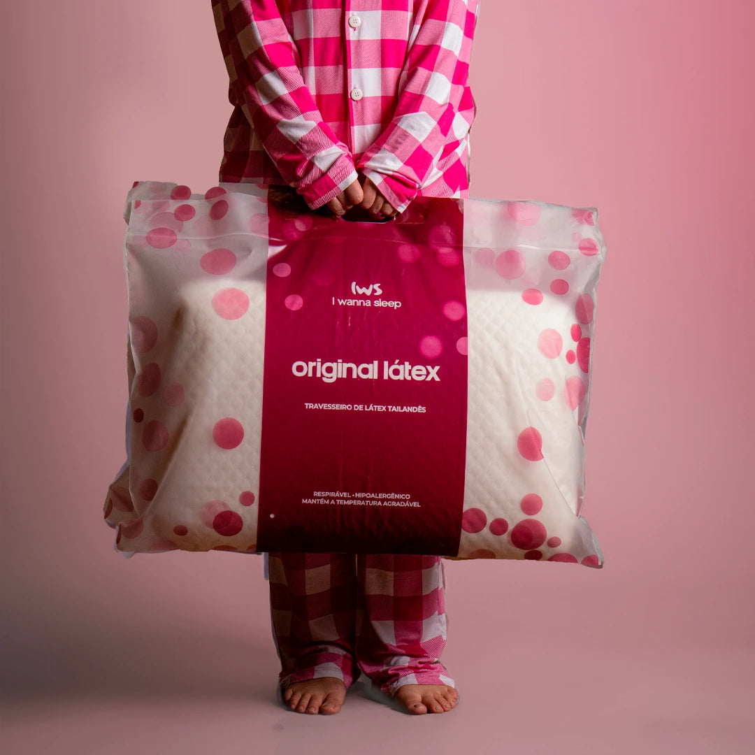 Mulher de pijama quadriculado nas cores rosa e branco segurando a embalagem linda do Travesseiro IWS Original Látex® na cor rosa ao centro, com transparências e bolinhas rosa do travesseiro
