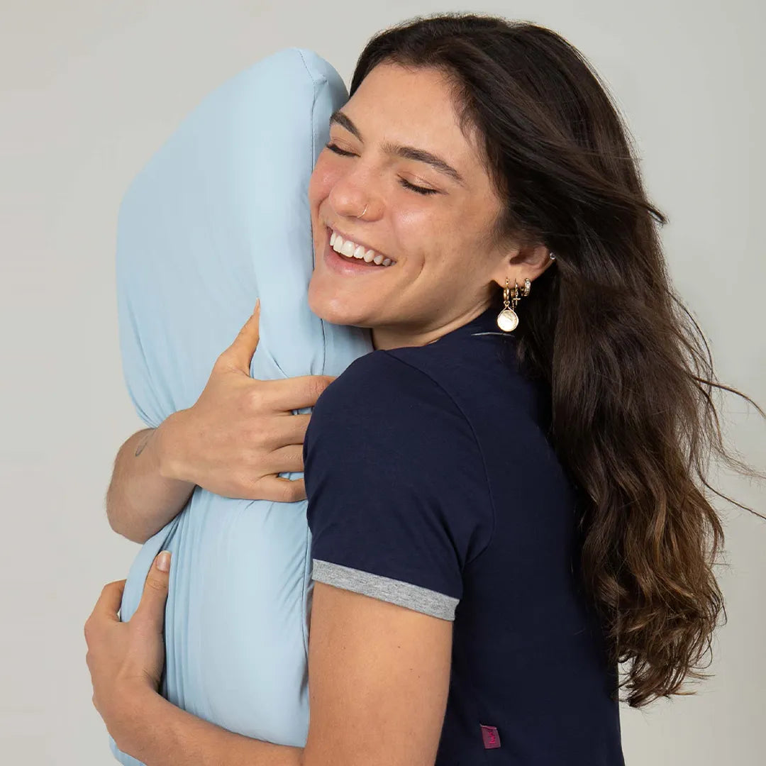 Mulher morena sorridente de pijama azul marinho abraçando o travesseiro em látex com sua capa refrescante, combina elasticidade e refrescância para um ajuste perfeito, proporcionando uma sensação suave para complementar sua experiência de sono, ideal para se manter refrescado em dias quente.