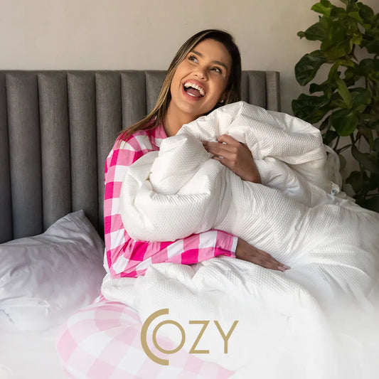 Mulher loira de pijama xadrez branco e rosa da i wanna sleep abraçando nosso cobertor quentinho Cozy com sua textura 3D Dots em sua cama que possi a cabeceira cinza. Do lado direito temos uma planta decorativa e na parte inferior da imagem temos a logo do produto sobrepondo a imagem
