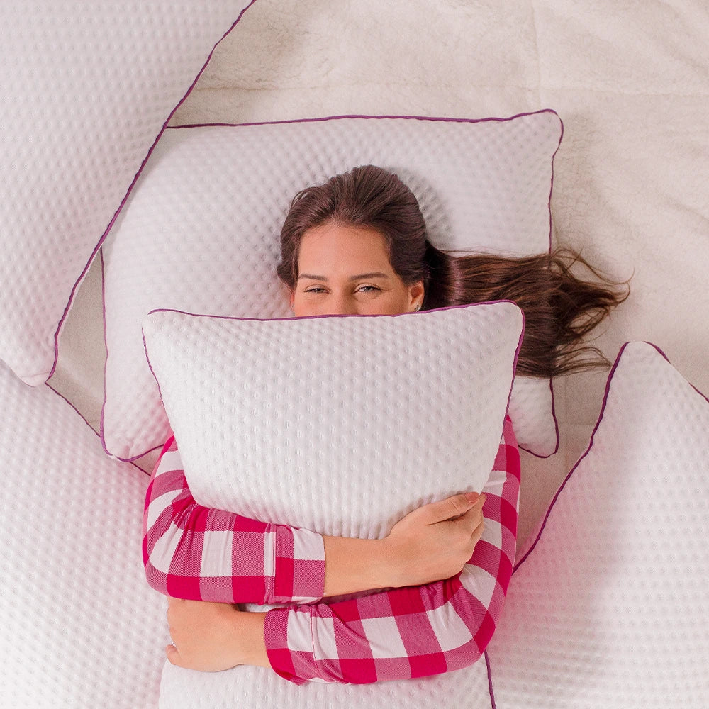 Mulher deitada de pijama quadriculado branco e rosa entre seus travesseiros confortáveis deli, abraçando um deles sobre o corpo deixando apenas os olhos de fora, testa e cabelo, fundo branco.