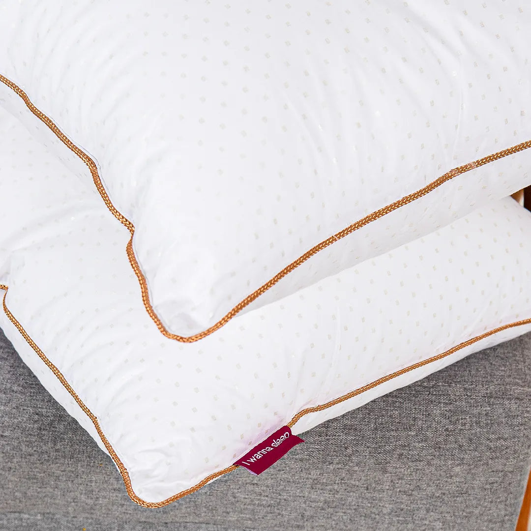 dois travesseiros de hotel Magnum pluma airfyber da cor branca com bordas douradas que trazem mais sofisticação ao seu design premium com fundo cinza.