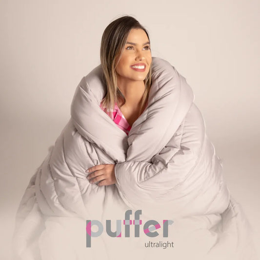 Mulher loira abraçada com o cobertor puffer da i wanna sleep, quentinho e macio para noites mais confortáveis. Na parte inferior temos a logo do produto Puffer ultralight sobrepondo a imagem