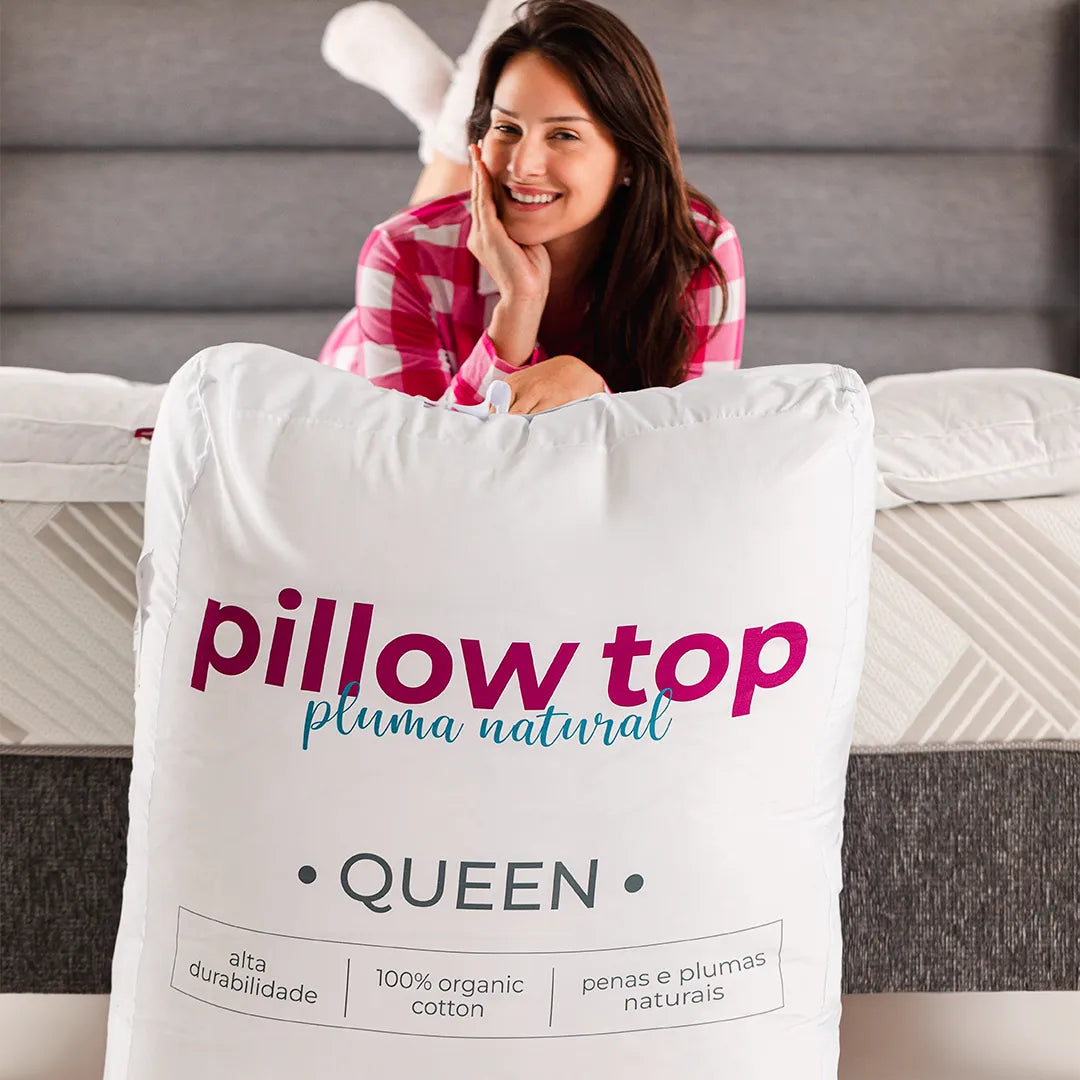 Mulher morena sorrindo de pijama xadrez branco e rosa deitada no colchão e apoiada na embalagem do pillow top iws pluma natural de ganso feita de tecido exclusivo para facilitar o armazenamento e preservar a qualidade do produto por mais tempo.
