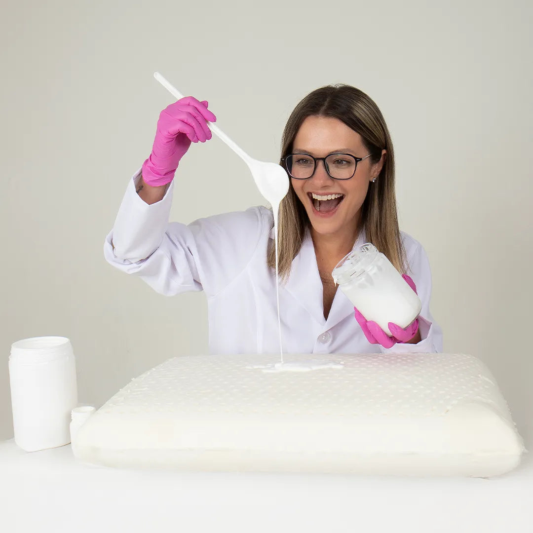 Mulher de óculos sorrindo manuseando com luvas a borracha liquida utilizada para fazer o travesseiro em látex. O travesseiro blue látex possui em sua matéria prima vinda diretamente das seringueiras da Tailândia, sendo 100% natural. A flexibilidade do material e resistência da borracha, torna o travesseiro adaptável às necessidades de todos os tipos de pessoas, atendendo à diferentes tipos de conforto.