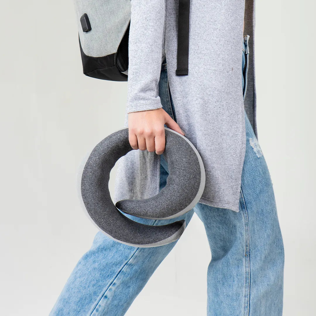 Mulher de calça jeans e blusa cinza caminhando com mochila nas costas, carregando seu travesseiro de pescoço Travel Pillow U - IWS Flow, ideal para viagens, garantindo descanso e conforto