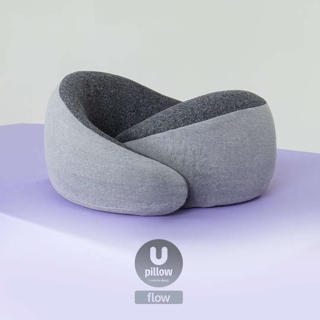 Travesseiro de pescoço Flow da I Wanna Sleep, na cor cinza, apoiado sobre uma superfície lilás com fundo branco, com o logo do produto na parte inferior da imagem.