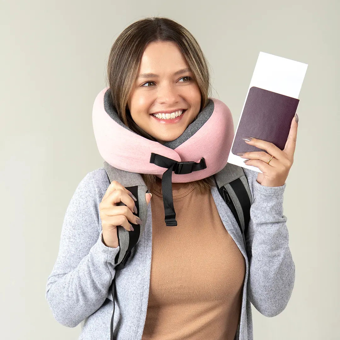 Mulher sorrindo, com travesseiro de pescoço Flow da IWS, mochila nas costas e passaporte na mão. Usando moletom cinza e blusa bege, pronta para uma viagem confortável e tranquila.