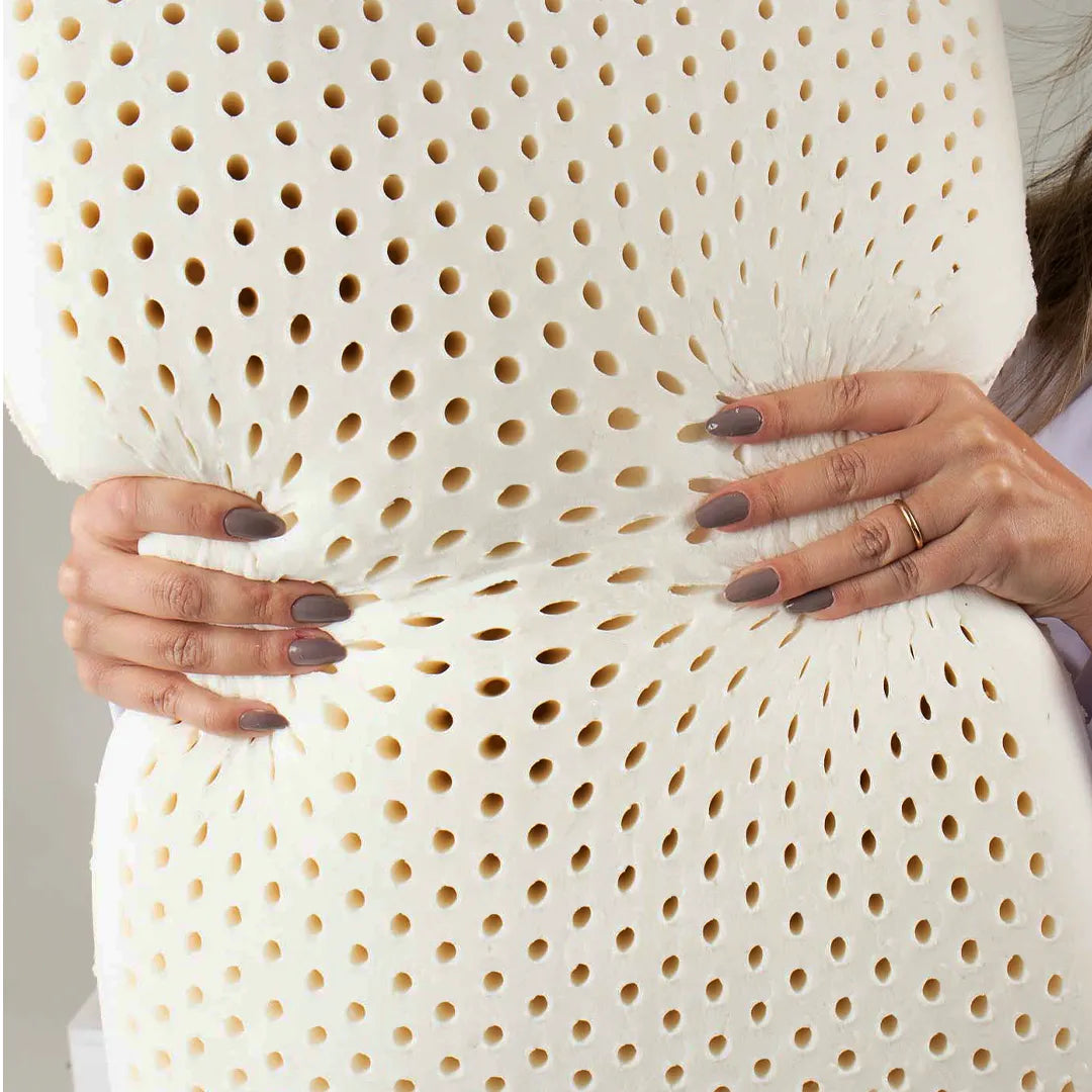 Travesseiro em látex Blue Látex IWS sendo pressionado por mãos femininas destacando seus centenas de furos que formam canais internos de circulação de ar e facilitam a respiração e ventilação do travesseiro, além de permitir evaporação rápida de umidade e transpiração, prevenindo odores e contaminações por ácaros, fungos e bactérias.