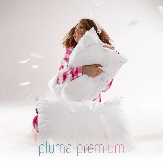 Mulher negra de pijama xadrex rosa e branco sorrindo enquanto abraça seu travesseiro de plumas de ganso premium que mantém sua maciez e forma original mesmo após longo uso, oferecendo conforto duradouro sem perder o suporte ideal. Fundo branco e logo do produto na parte inferior da imagem com penas de ganso voando
