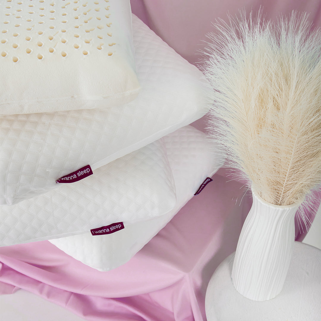 Travesseiros IWS Original Látex® empilhados com a logo da i wanna sleep em evidência, fundo rosa e um vaso de planta branco ao lado