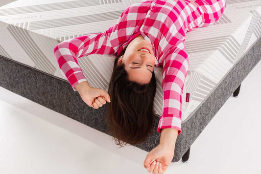 Mulher jovem sorrindo de felicidade enquanto relaxa de costas em um colchão IWS de última geração, vestindo um confortável pijama xadrez rosa e branco.