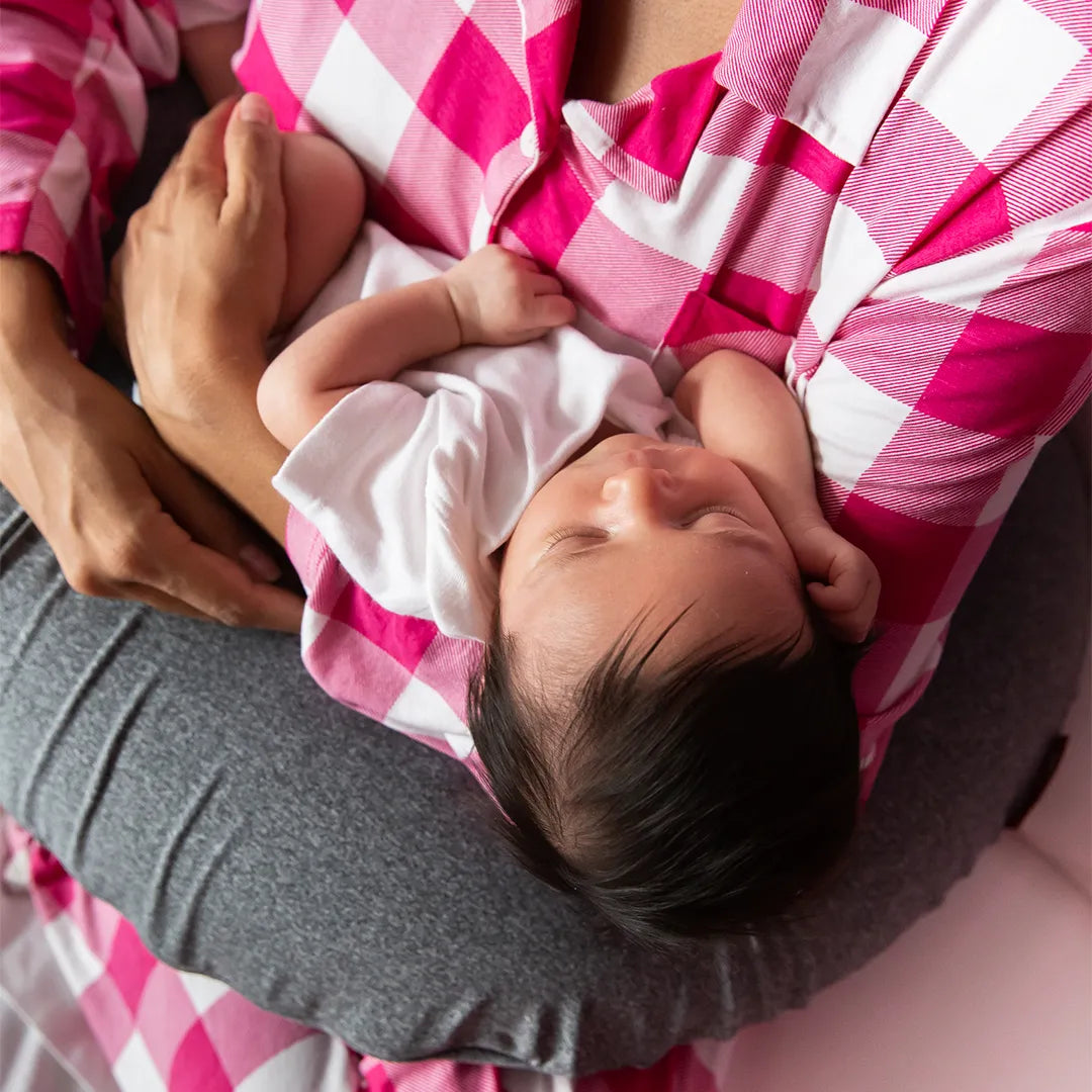 Mãe com pijama xadrez rosa e branco segurando seu bebê nos braços com o apoio do travesseiro de amamentação Nest da IWS que possui o ajuste perfeito para mãe e bebê, garantindo pega correta e alívio de dores