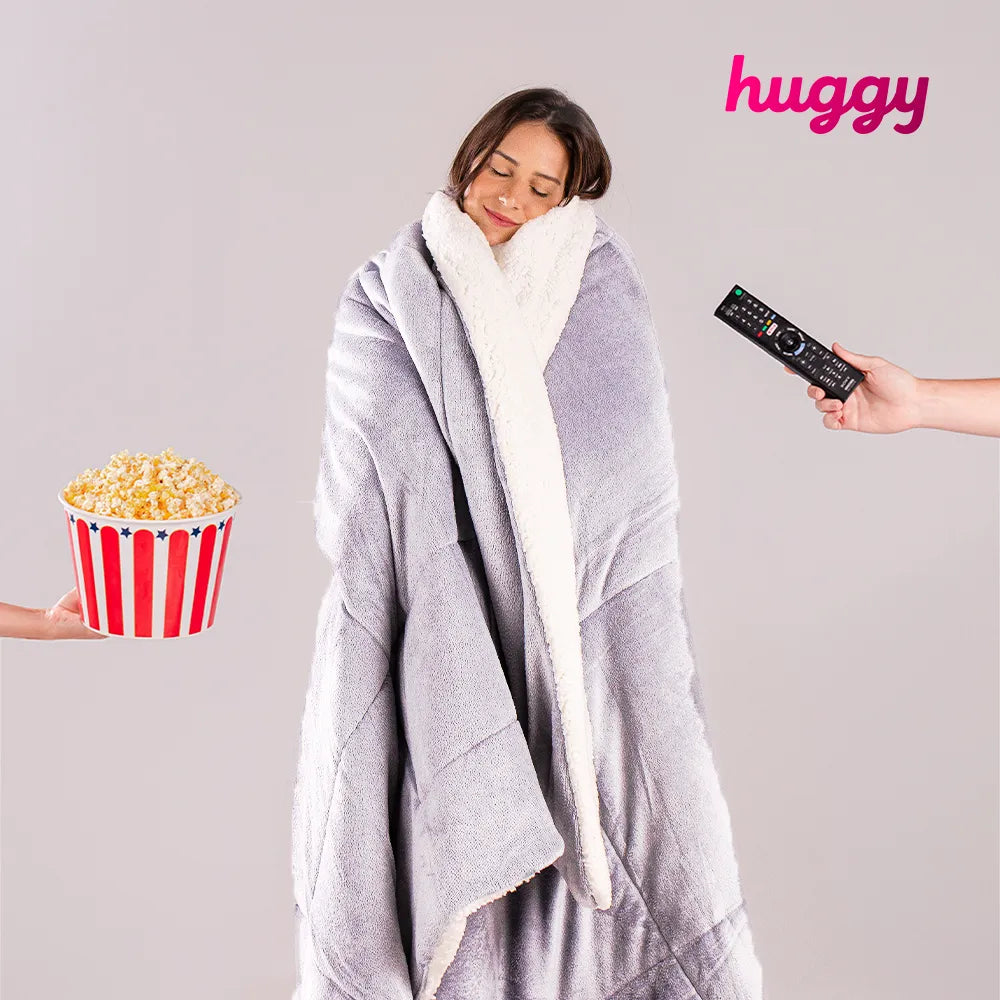 Mulher morena envolvida com seu cobertor quentinho Huggy cinza coala sendo oferecido do seu lado direito um controle de tv e do esquerdo uma pipoca, perfeito para dias frios em que queremos assistir um filme no maior conforto possível.