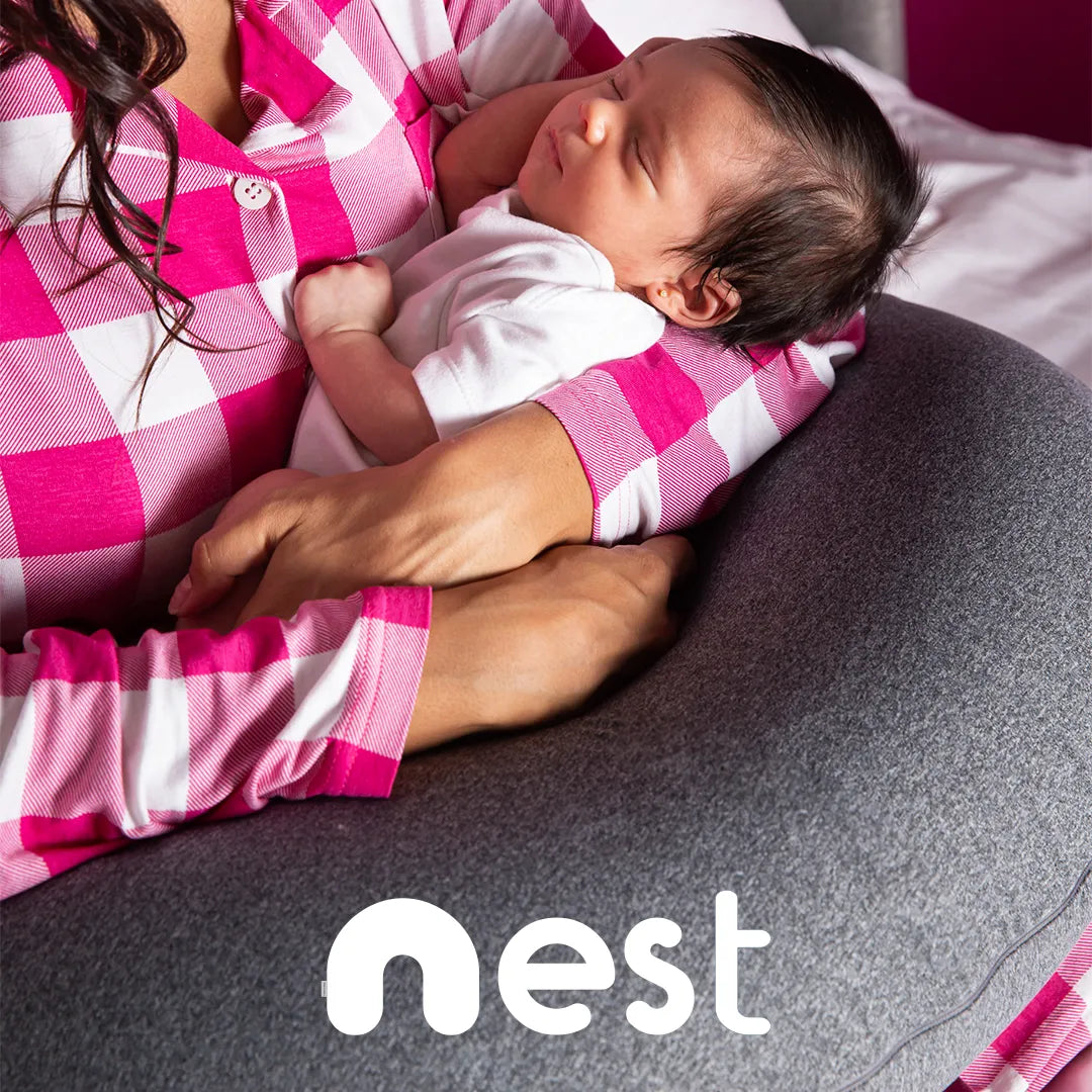 Mãe morena de pijama xadrez rosa e branco segurando seu bebê no colo com o apoio da almofada de amamentação confortável Nest da I wanna sleep, na parte inferior da imagens temos a logo da almofada Nest em destaque 