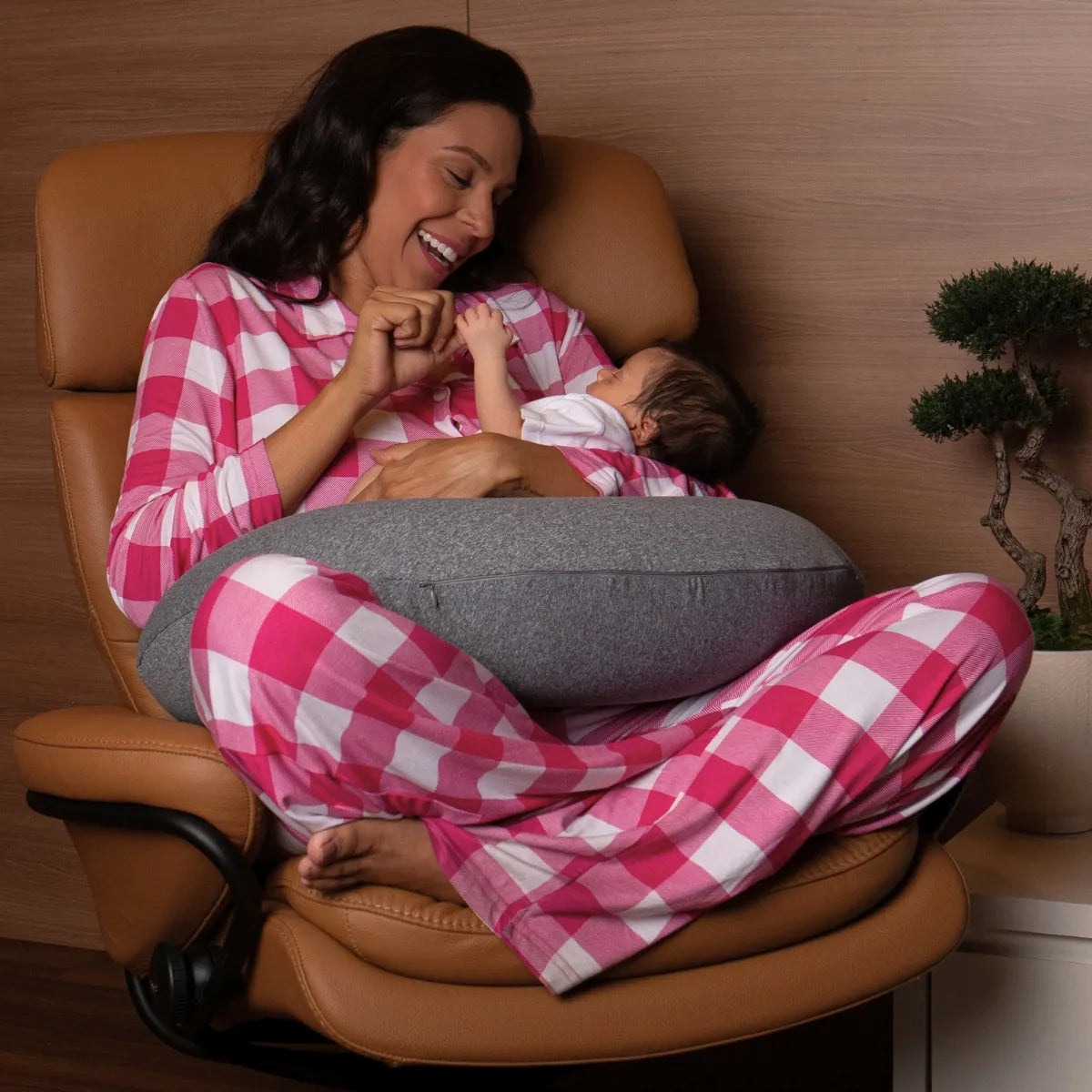 Mãe segurando e brincando com seu bebê utilizando como apoio confortável a almofada de amamentação nest da I wanna sleep, sentada em uma poltrona cor caramelo com fundo em madeira e um caso de planta ao seu lado.