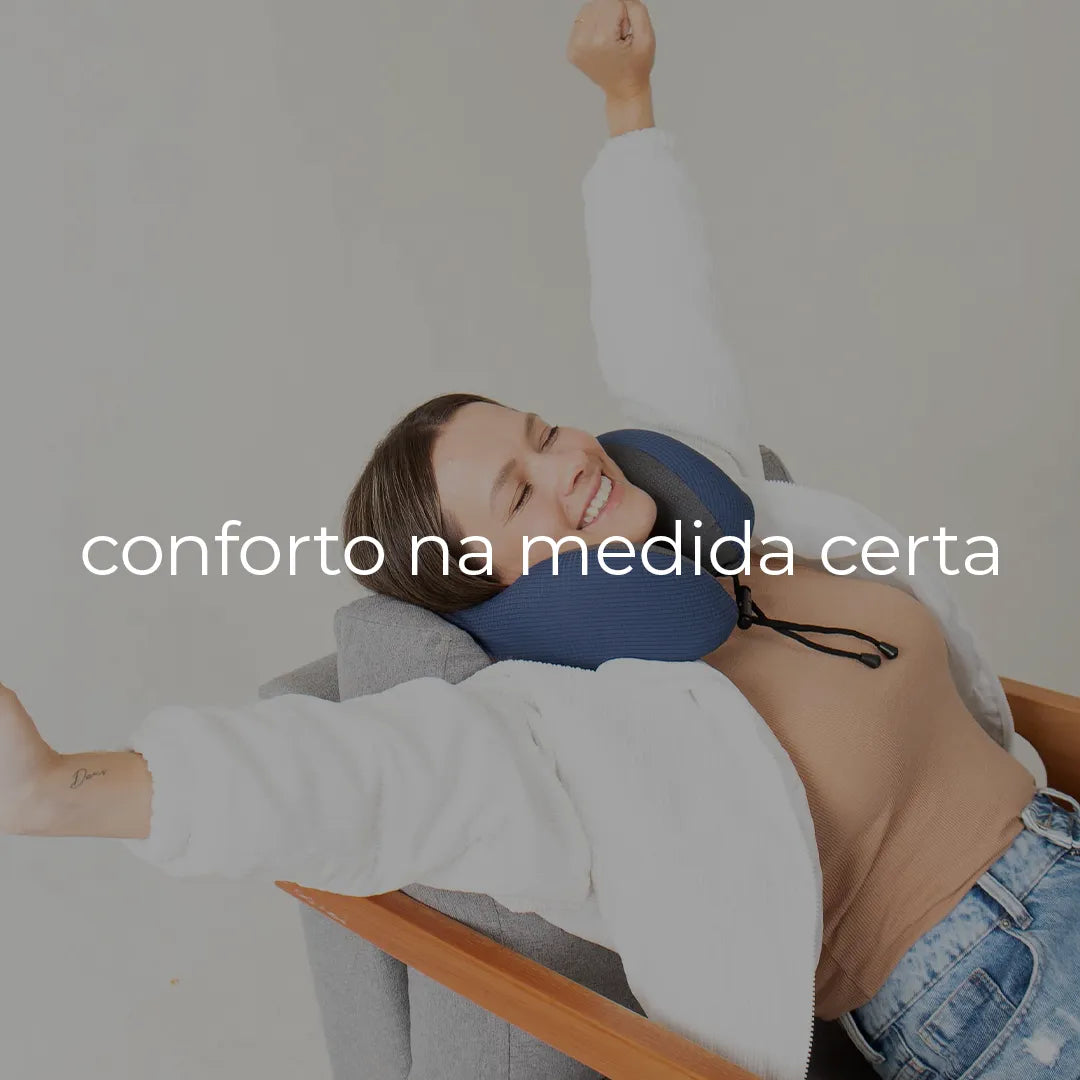 Mulher loira sorrindo e se espreguiçando em uma cadeira cinza com braços de madeira, confortável com seu travesseiro de pescoço da I Wanna Sleep. Veste jaqueta branca, blusa nude e calça jeans.