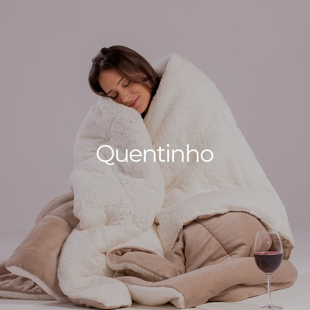 Mulher morena contente e abraçada de forma aconchegante na sua coberta quentinha Huggy, perfeito para dias de descanso e relaxamento, representando isso temos uma taça de vinho no chão ao lado da modelo. Temos a palavra "quentinho" sobrepondo a imagem