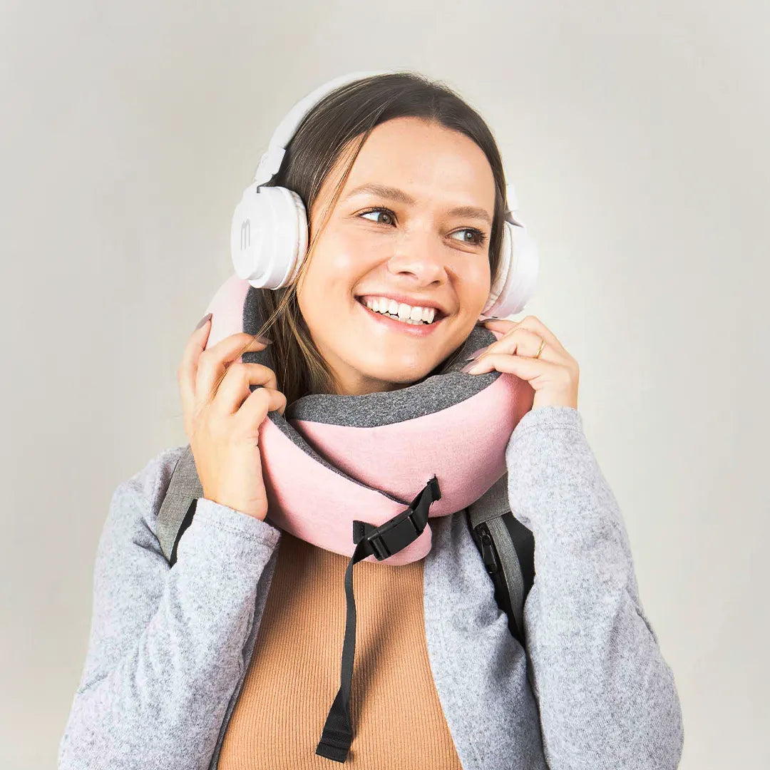 Mulher sorrindo, apoiando as mãos no travesseiro de pescoço para viagem. Usa moletom cinza, mochila nas costas e fones de ouvido brancos, pronta para uma viagem confortável.