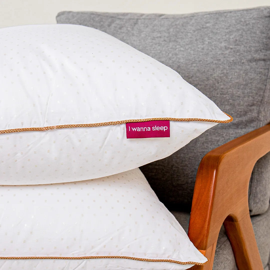 Dois travesseiros de hotel Magnum Pluma IWS empilhados em cima de uma poltrona cinza com braços de madeira. Imaginado para alcançar até 15 cm de pura maciez, este travesseiro é um sinônimo de conforto.